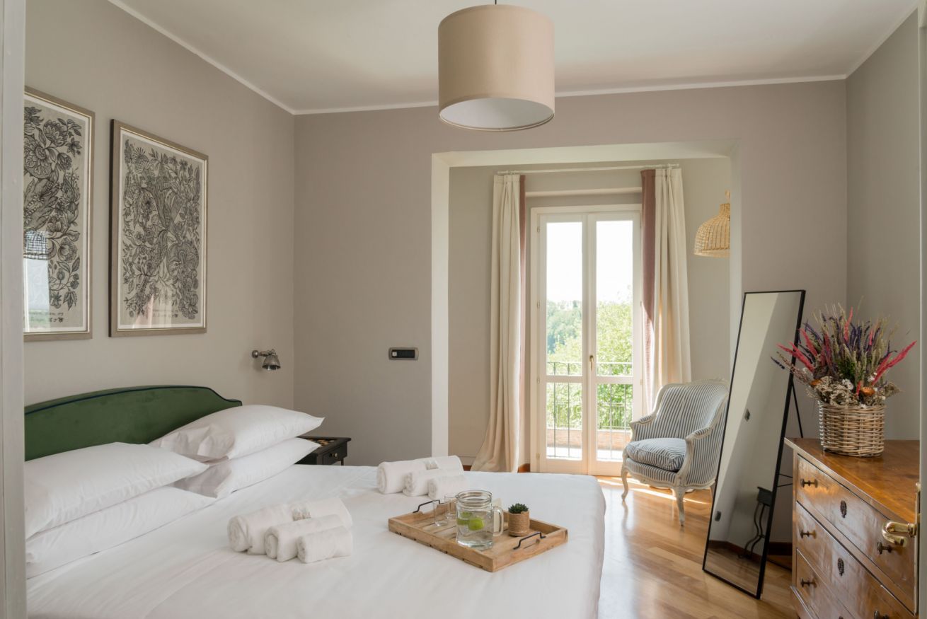 A neatly made bed with white linens, a bedside table with a lamp, a chair, a mirror, and artwork on the walls.