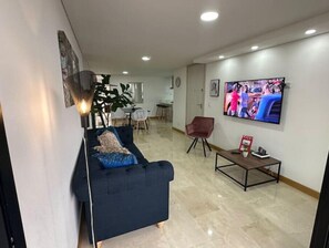 Ein Wohnzimmer mit einer Couch, einem Couchtisch und einem Fernseher. Im Hintergrund ist ein Essbereich zu sehen.