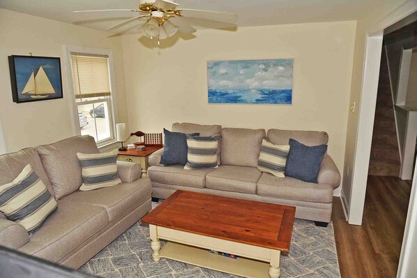 A living room with a beige sofa set, a wooden coffee table, a ceiling fan, and two paintings on the walls.