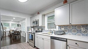 A modern kitchen with white cabinetry, a microwave, an oven, and a dishwasher. There is a dining area with a table and chairs visible in the background. - Let the Good Times Roll/ Spacious Retreat 4BE 3BA (Atlanta)