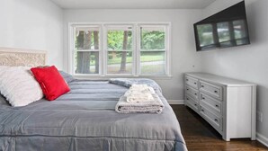 A bedroom with a bed, a dresser, a TV mounted on the wall, and a window with blinds. - Let the Good Times Roll/ Spacious Retreat 4BE 3BA (Atlanta)
