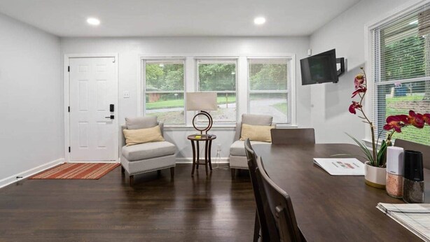 A room with a dark wooden floor, white walls, a white door, two windows with blinds, a TV mounted on the wall, a table with chairs, and a small side table with a lamp. - Let the Good Times Roll/ Spacious Retreat 4BE 3BA (Atlanta)