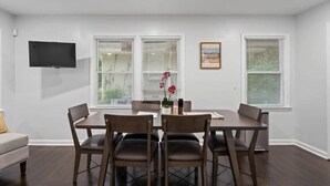 A dining area with a table and chairs, a flat-screen TV mounted on the wall, and three windows with blinds. - Let the Good Times Roll/ Spacious Retreat 4BE 3BA (Atlanta)