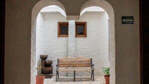 A bench in a corridor with white walls and stone flooring, flanked by two potted plants and a sign indicating an evacuation route. - Hotel Real del Carmen (San Cristóbal de las Casas)