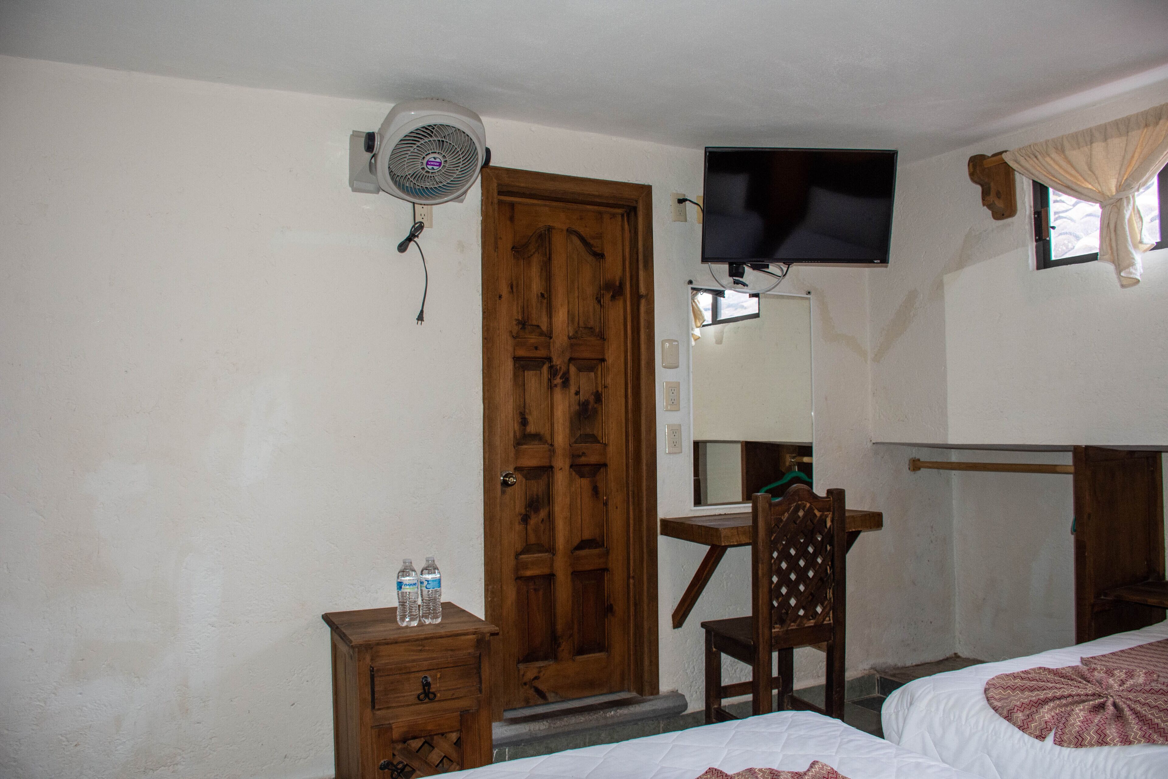 A room with a wooden door, a desk with a chair, a bed, a television mounted on the wall, and a fan.