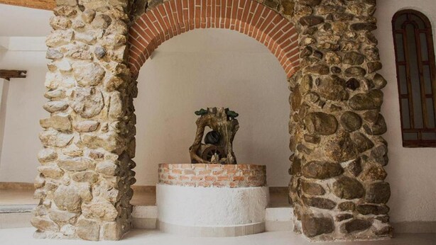 A stone archway with a rustic stone pillar on the left and a stone wall on the right, leading to a circular stone platform with a decorative tree trunk sculpture in the center. - Hotel Real del Carmen (San Cristóbal de las Casas)