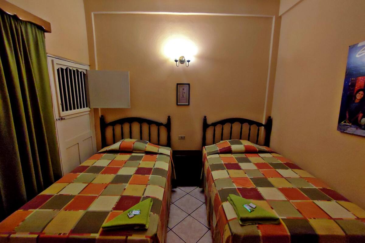 Une chambre avec deux lits simples, une fenêtre avec des stores, un luminaire fixé au mur et un tableau encadré.