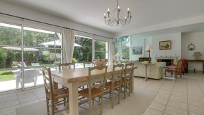 A spacious dining area with a large wooden table and chairs, a chandelier, and large windows offering a view of the garden. - Hossegor Proche Lac, Villa 8 Personnes, Piscine (HOSSEGOR)