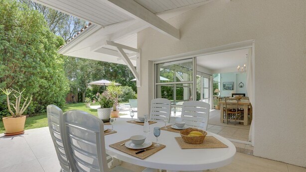 A white table set outdoors with chairs, a glass door leading to a kitchen, and potted plants. - Hossegor Proche Lac, Villa 8 Personnes, Piscine (HOSSEGOR)