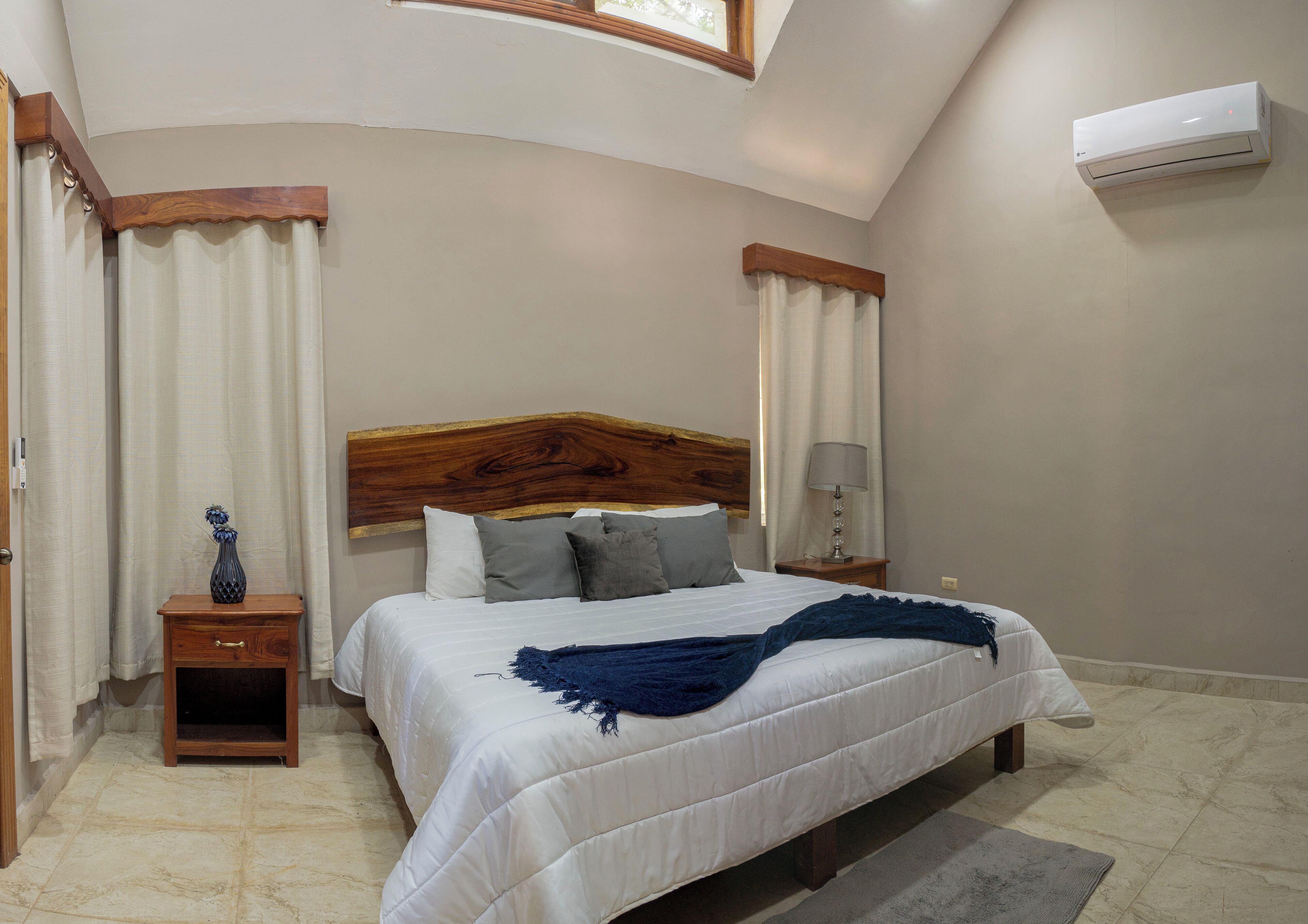 Un dormitorio con cabecera de madera, una mesita de noche con lámpara, un jarrón y una cama con una manta azul.