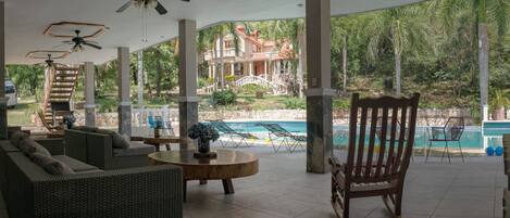 A covered outdoor area with wicker furniture, a pool, and a view of a large house with a bridge.