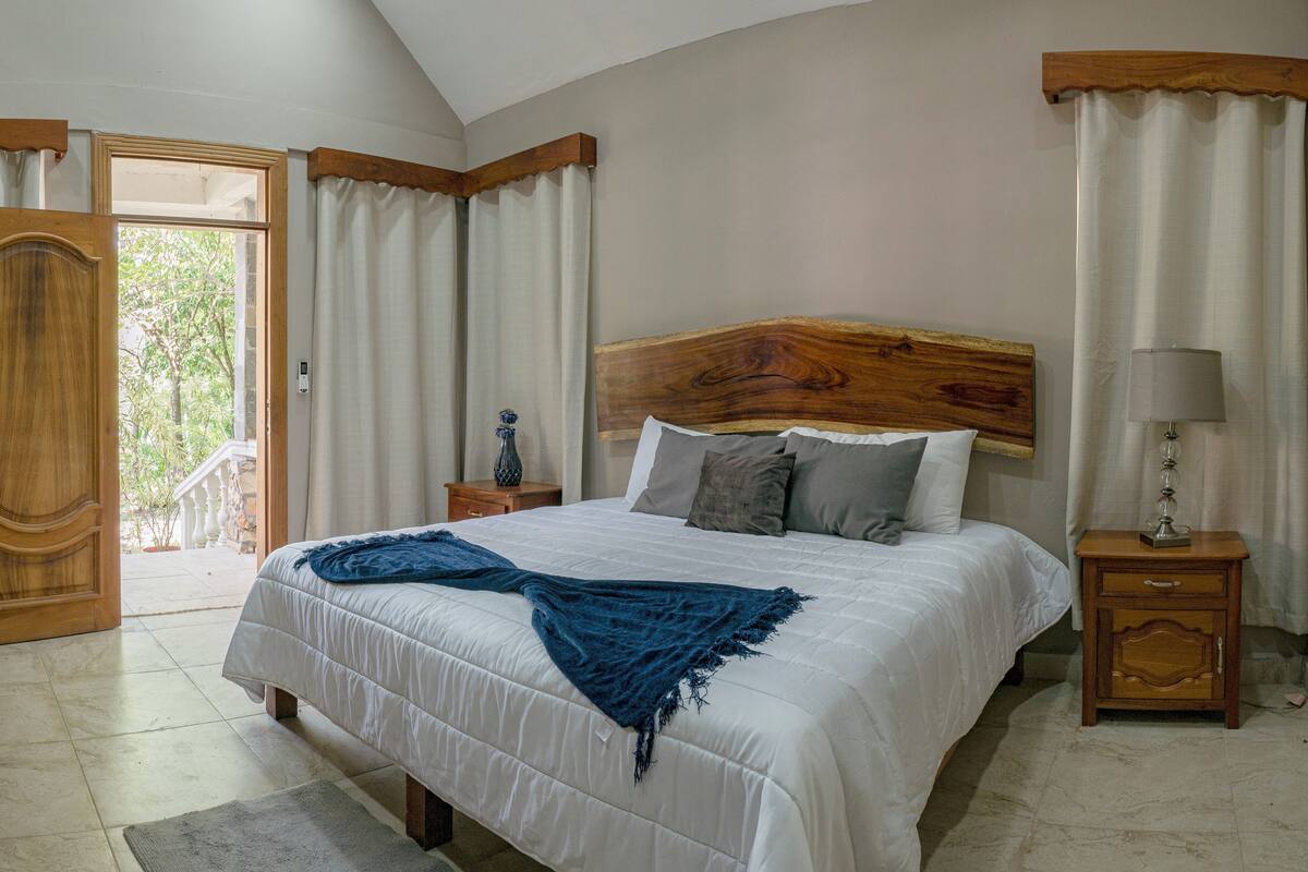 Une chambre avec un grand lit, une tête de lit en bois, une table de chevet avec une lampe, et une vue sur l’extérieur par une porte ouverte.