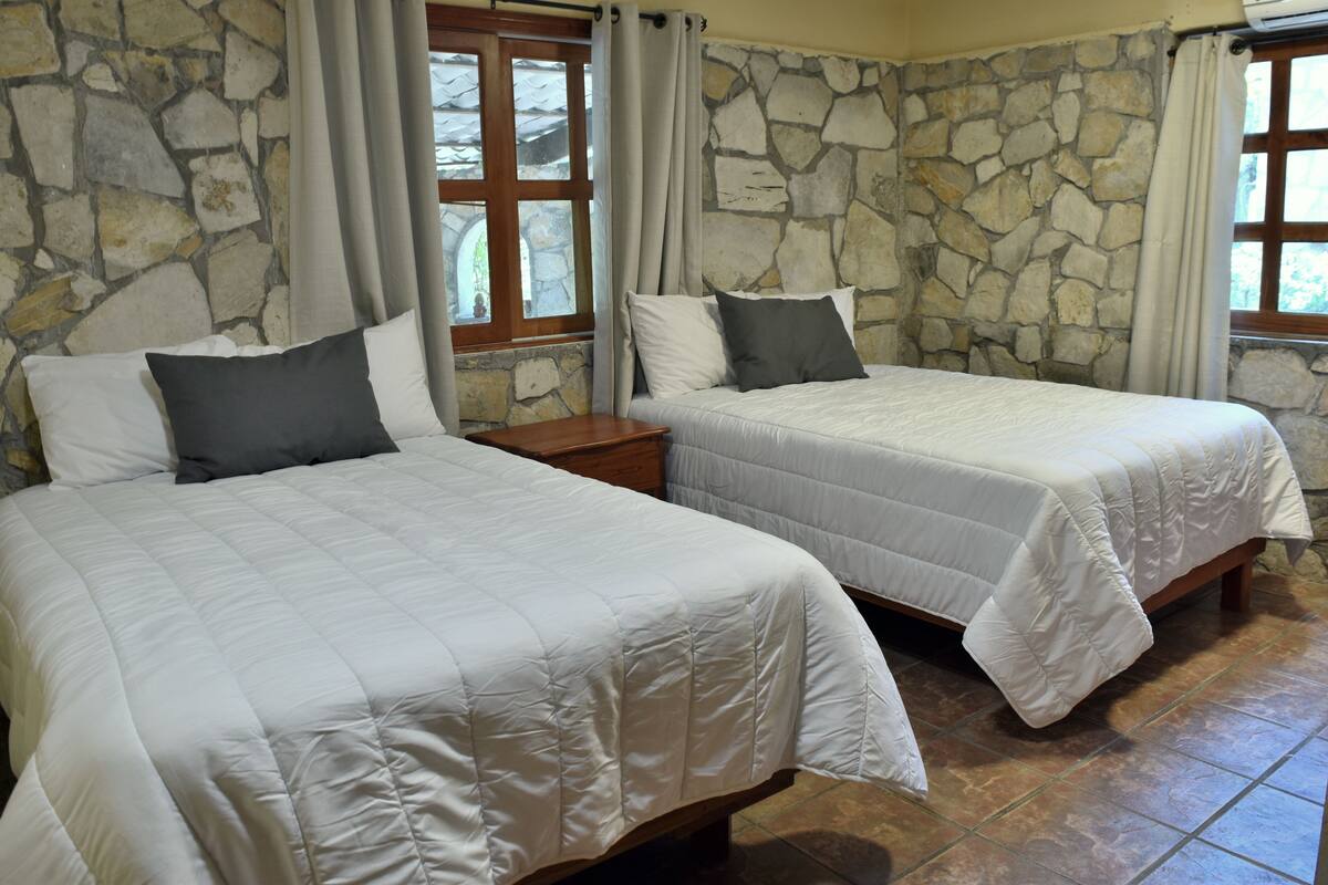 Deux lits avec du linge de lit blanc et des oreillers gris, un mur en pierre et une fenêtre avec des rideaux.