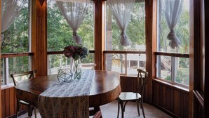 Une table à manger en bois, recouverte d’une nappe à motifs, entourée de chaises, dans une pièce dotée de grandes fenêtres et de rideaux.