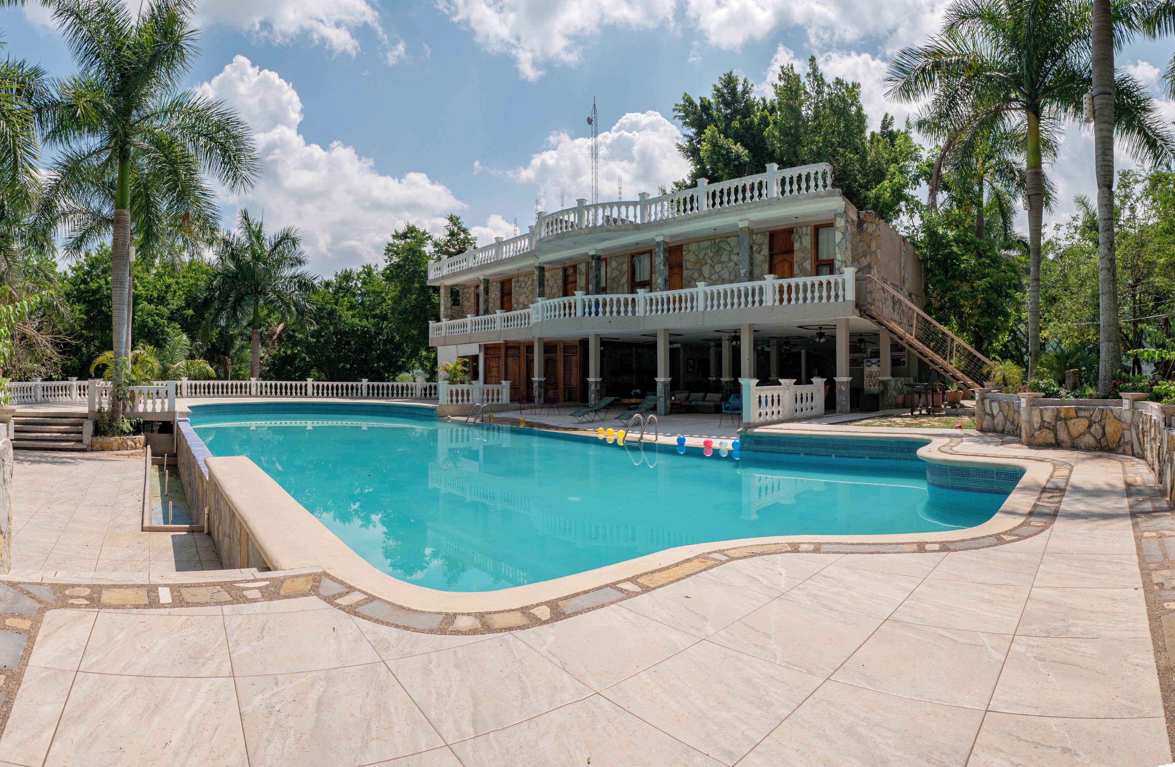 Una amplia piscina con plataforma de piedra, rodeada de palmeras y un edificio de dos pisos con balcones.