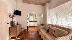 A spacious living room with a high ceiling, a wooden ceiling, a woven sofa, a wooden coffee table, and a kitchen visible through an archway. - Cosy cottage near the beautiful West Coast beaches (Holetown)