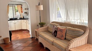 A cozy living room with a woven sofa, a small wooden table, and a fan. A kitchen with white cabinets and a tiled floor is visible through an archway. - Cosy cottage near the beautiful West Coast beaches (Holetown)