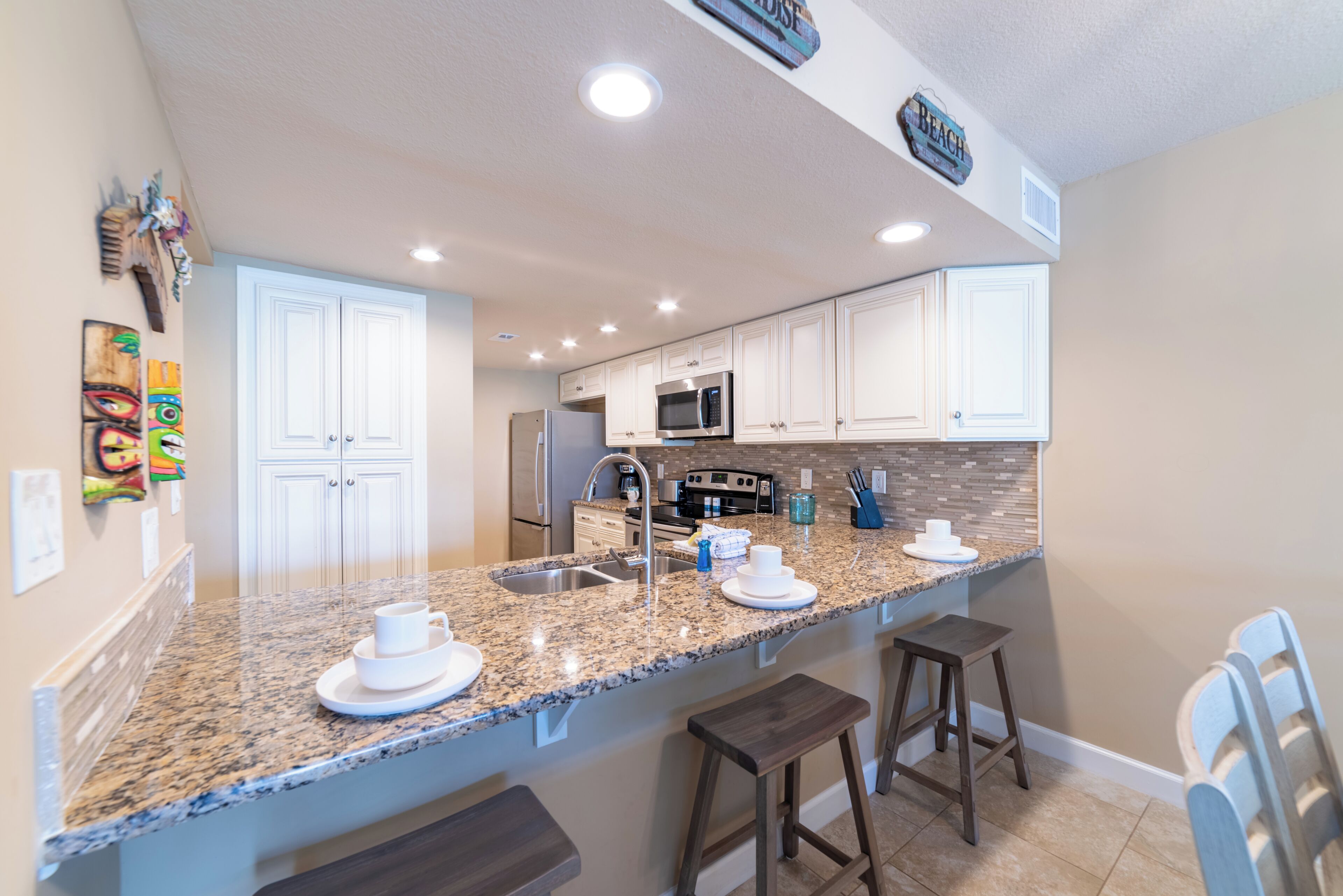 A modern kitchen with granite countertops, white cabinets, and stainless steel appliances. There are two bar stools and a built-in island with a sink.