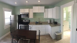 A compact kitchen with white cabinets, a stainless steel refrigerator, a microwave, and a stove. There is a dining table with chairs, a trash can, and a window with blinds. - 19988 Front Beach Rd (Panama City Beach)