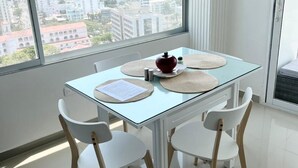 A modern dining area with a glass table and white chairs, a view of a cityscape through large windows, and a white column in the corner.