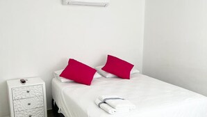 A neatly made bed with white bedding and two bright pink pillows, a white nightstand with drawers, and a white air conditioning unit on the wall.