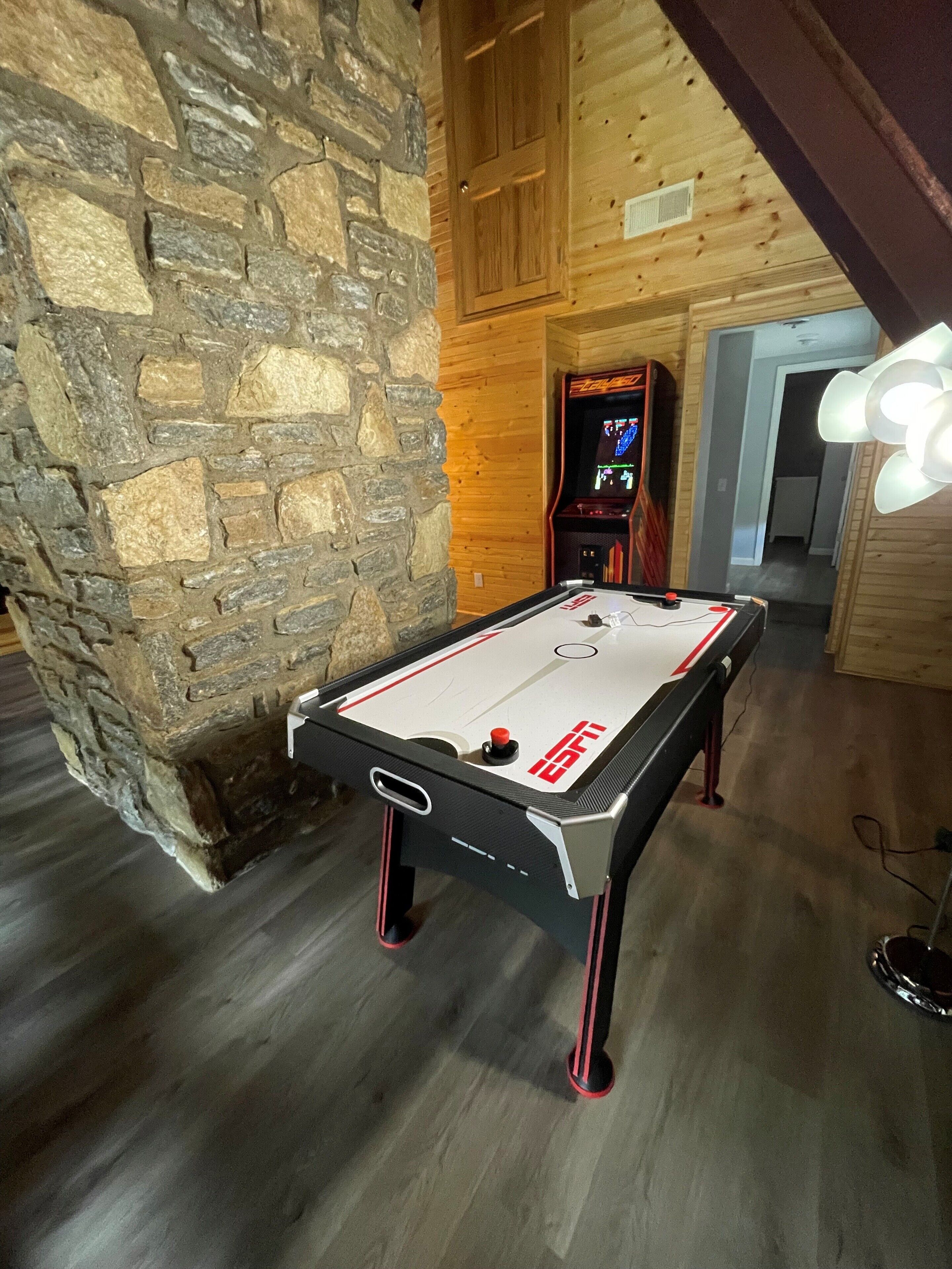 A pool table in a room with stone walls and wooden beams, a video game machine, and a ceiling light fixture.