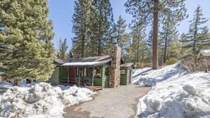 A cabin with a stone chimney, surrounded by snow and tall pine trees. - Village and Lake Escape - Hot Tub, Walk to Lake & Village, Barbecue (Big Bear Lake)