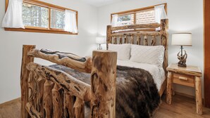 A rustic wooden bed with a unique design, a bedside table with a lamp, and a window with curtains. - Village and Lake Escape - Hot Tub, Walk to Lake & Village, Barbecue (Big Bear Lake)