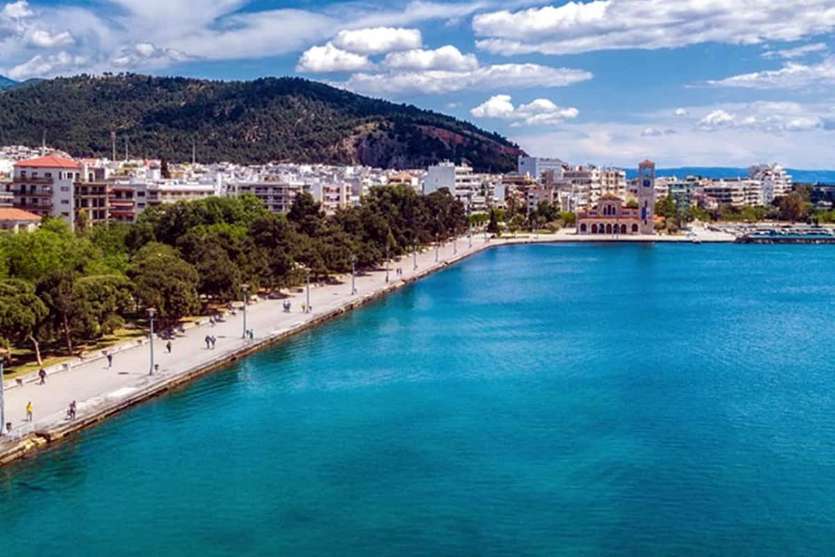 A coastal cityscape with a clear blue sea, a promenade lined with trees, and buildings in the background.