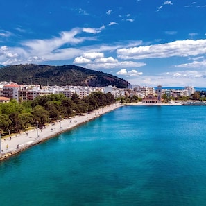 A coastal cityscape with a clear blue sea, a promenade lined with trees, and buildings in the background. - Volos Terra Deluxe Residences (Volos)