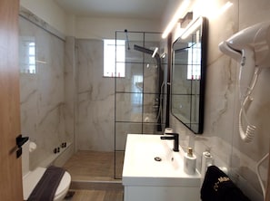 A modern bathroom with a glass shower enclosure, a white sink with a black faucet, a handheld hair dryer, and a towel on the floor. - Volos Terra Deluxe Residences (Volos)