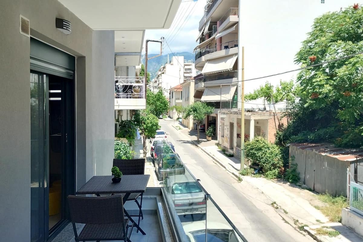 A balcony with a table and chairs, overlooking a street with parked cars and buildings.