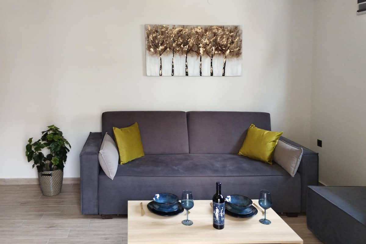 A modern living room with a grey sofa, two yellow cushions, a wooden coffee table with wine glasses and a bottle, and a wall-mounted abstract painting.