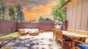 Ein Garten mit Holztisch und -stühlen, einer Feuerstelle und einem überdachten Grillbereich.