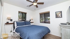 Ein Schlafzimmer mit einem Bett, einem Deckenventilator, zwei Fenstern, einem Schreibtisch mit Computer und einem Fernseher.