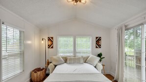 Une chambre avec un grand lit, deux fenêtres équipées de stores, une lampe sur pied et une vue sur l’extérieur.