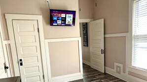 Un dormitorio con una televisión montada en la pared mostrando diferentes servicios de transmisión, una cama con edredón blanco y suelo de madera.