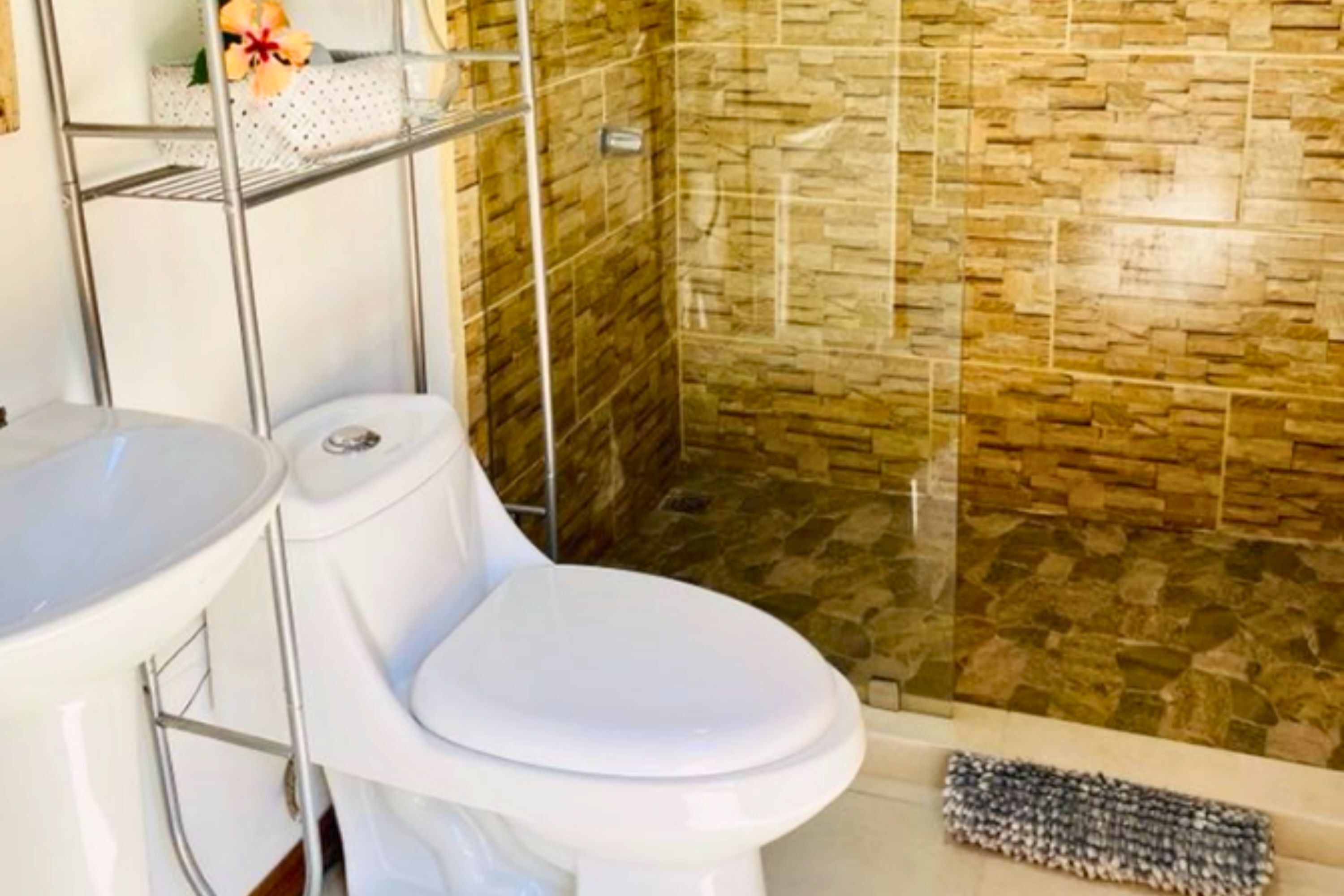 Une salle de bain avec des toilettes blanches, un lavabo blanc et un espace douche avec un mur en pierre.