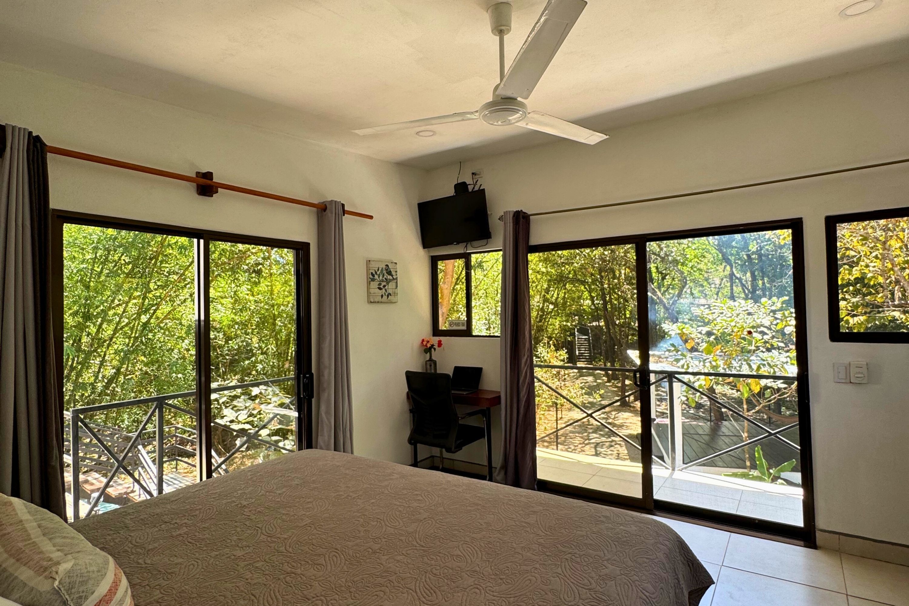 Quarto com cama, escrivaninha, cadeira, televisão e grandes portas de vidro deslizantes que dão para uma varanda com vista para o verde.