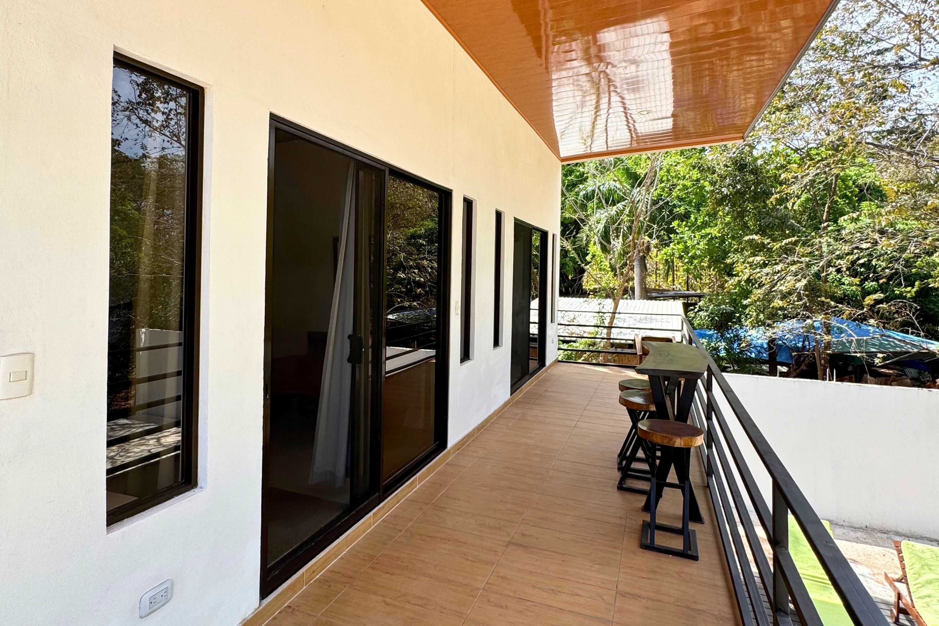 Un balcon extérieur abrité, doté d’un plancher en bois, de fenêtres à cadre noir et d’une table avec des chaises.