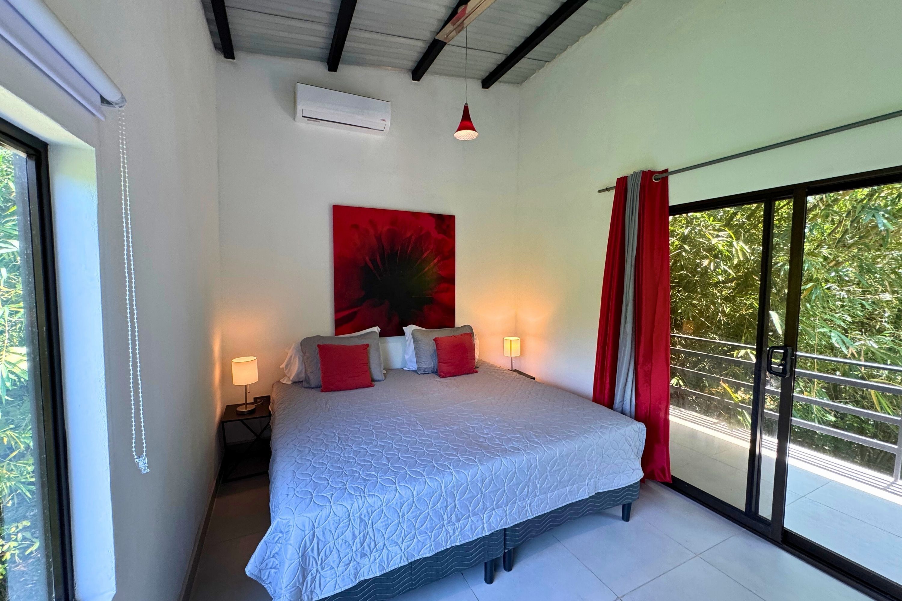 A bedroom with a large bed, a red abstract painting, a nightstand with a lamp, and a view of greenery through a sliding door.
