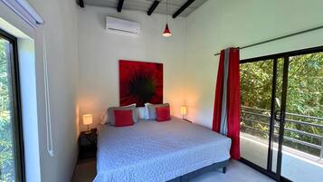 A bedroom with a large bed, a red abstract painting, a nightstand with a lamp, and a view of greenery through a sliding door.