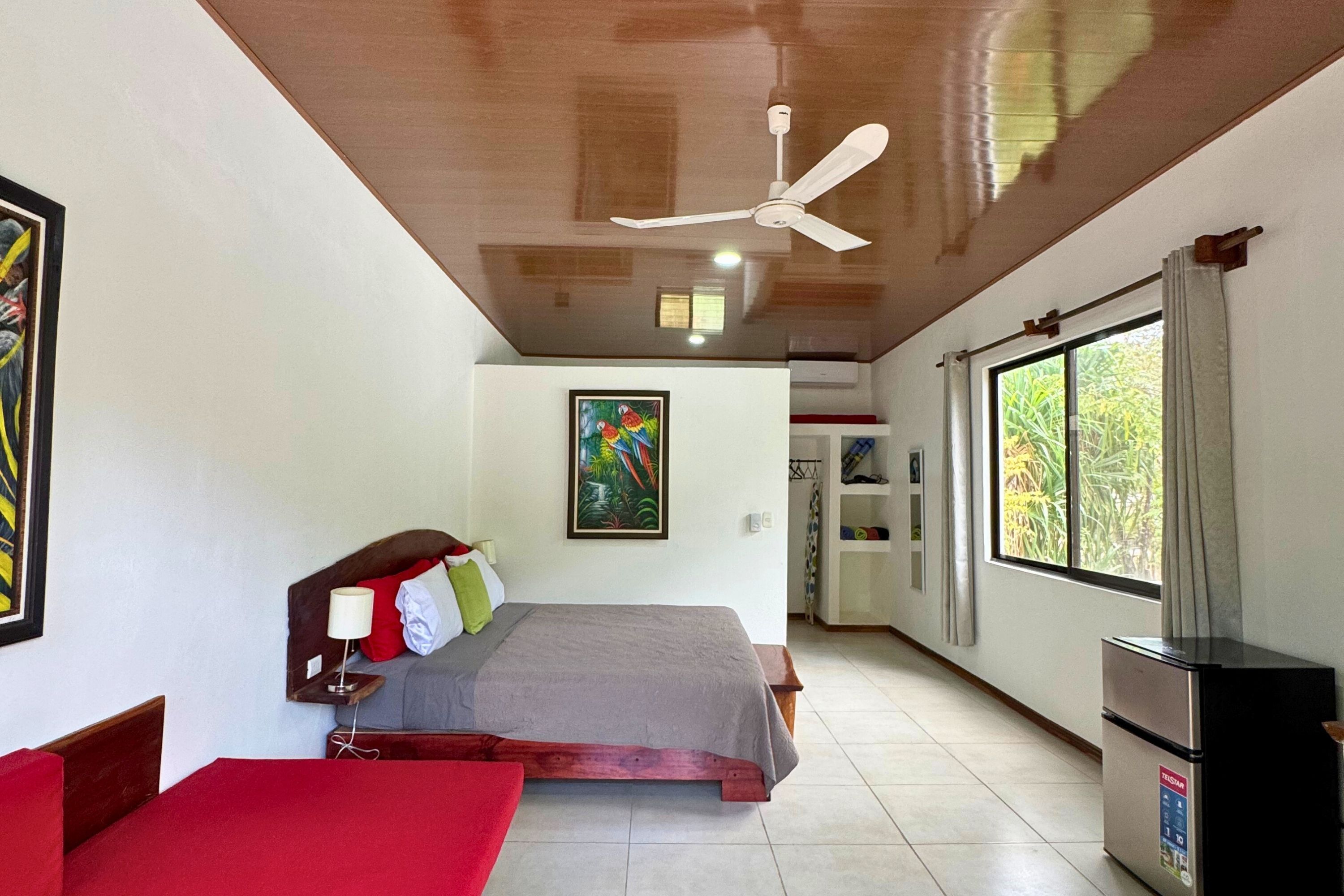 Habitación de hotel con cama, escritorio, refrigerador, ventilador de techo, ventana y un cuadro en la pared.