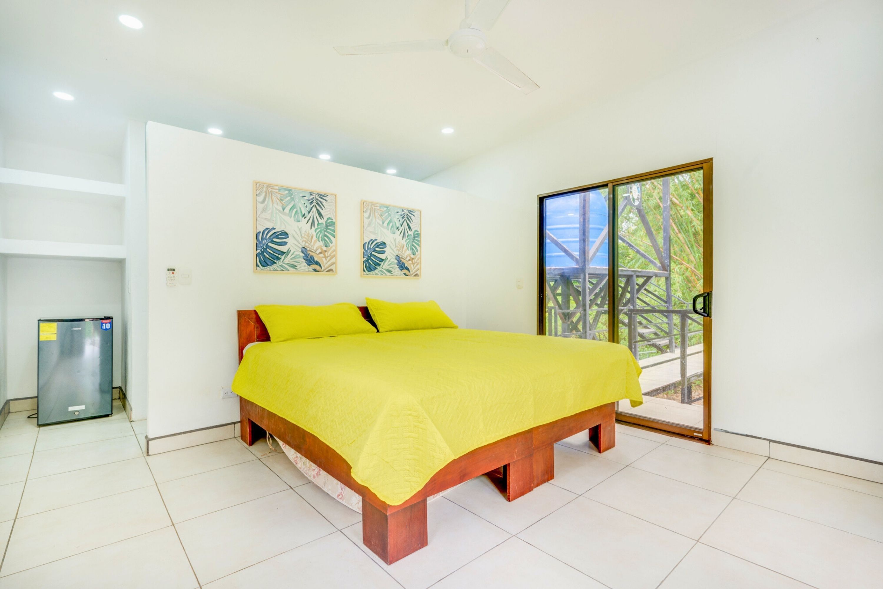 A bedroom with a bed, a ceiling fan, a refrigerator, and a sliding glass door to an outdoor area.