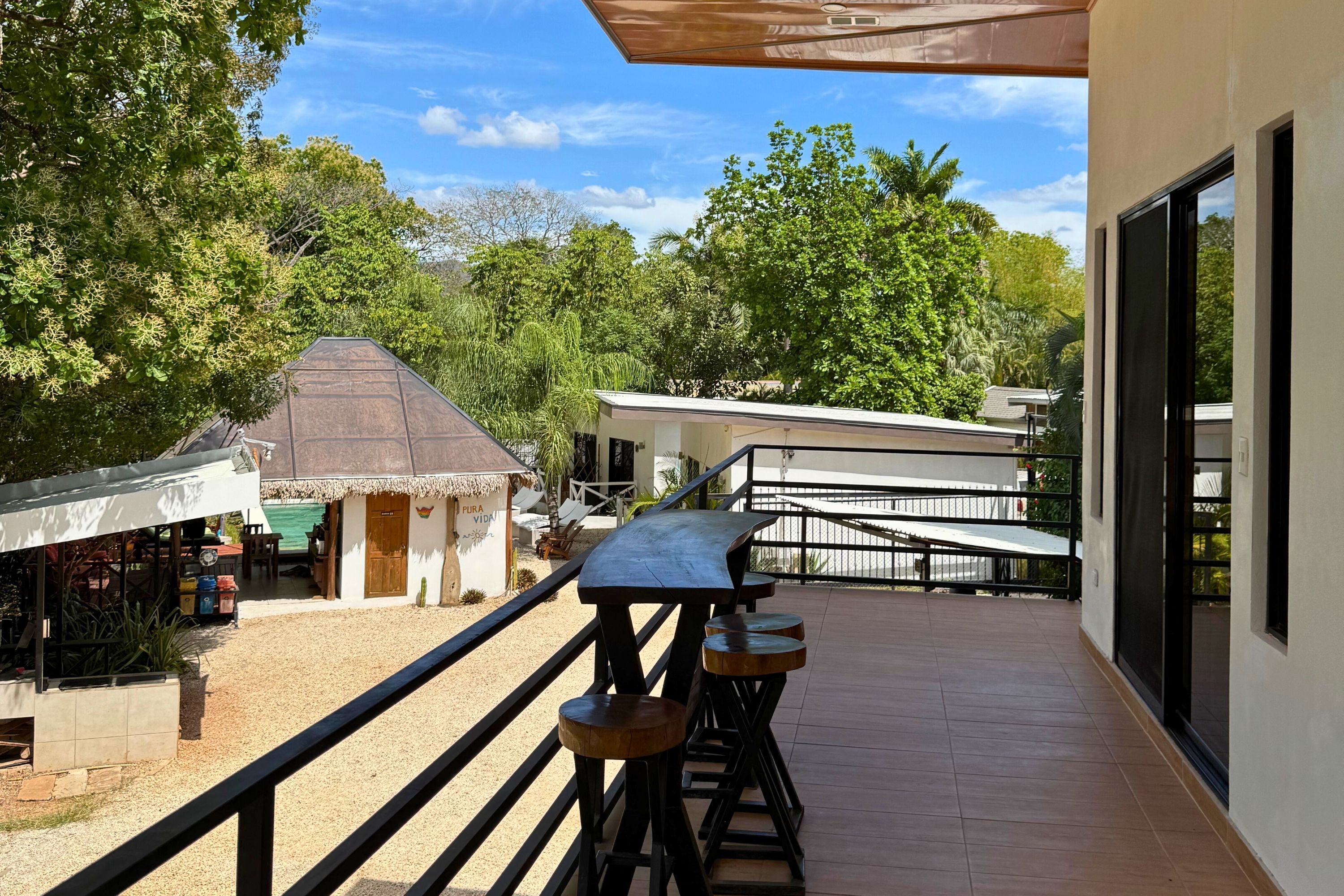 A balcony with a wooden table and stools, overlooking a restaurant and a building with a thatched roof.