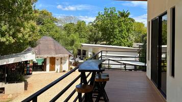 A balcony with a wooden table and stools, overlooking a restaurant and a building with a thatched roof.