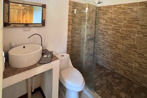 A bathroom with a stone wall, a white sink, a toilet, and a glass shower enclosure. - Antema Lodge (Tamarindo)