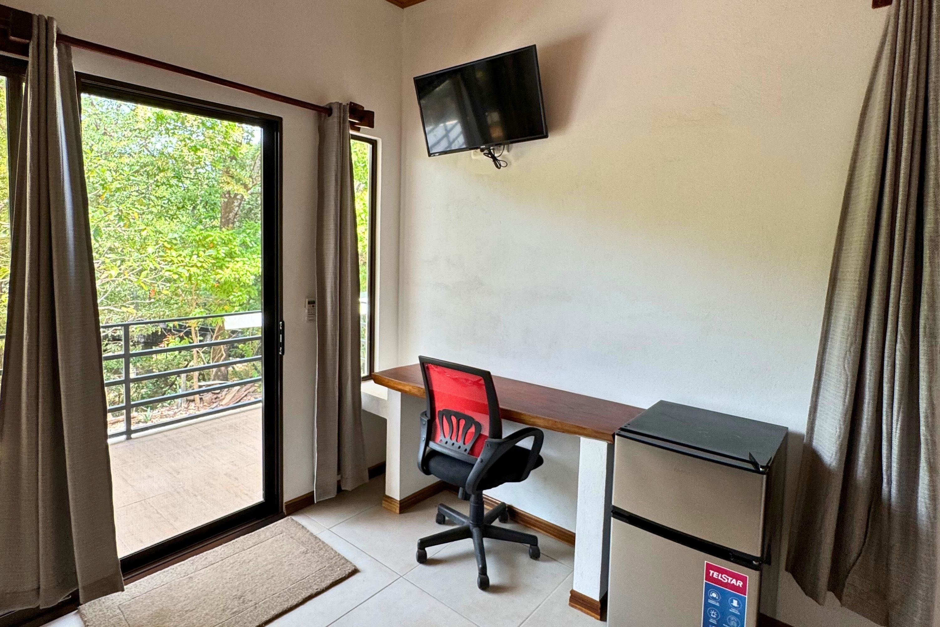 A room with a desk, chair, TV, and a view of greenery outside.
