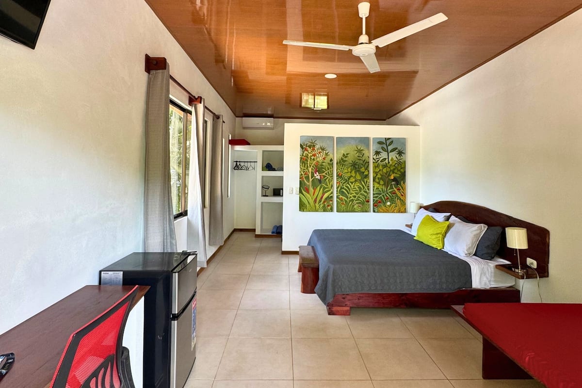 A bedroom with a bed, a desk, a ceiling fan, and a window with a view of greenery.