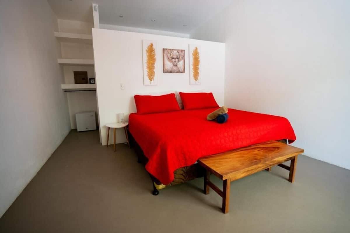 A bedroom with a red bedspread, a wooden bench, and two wall-mounted artworks.